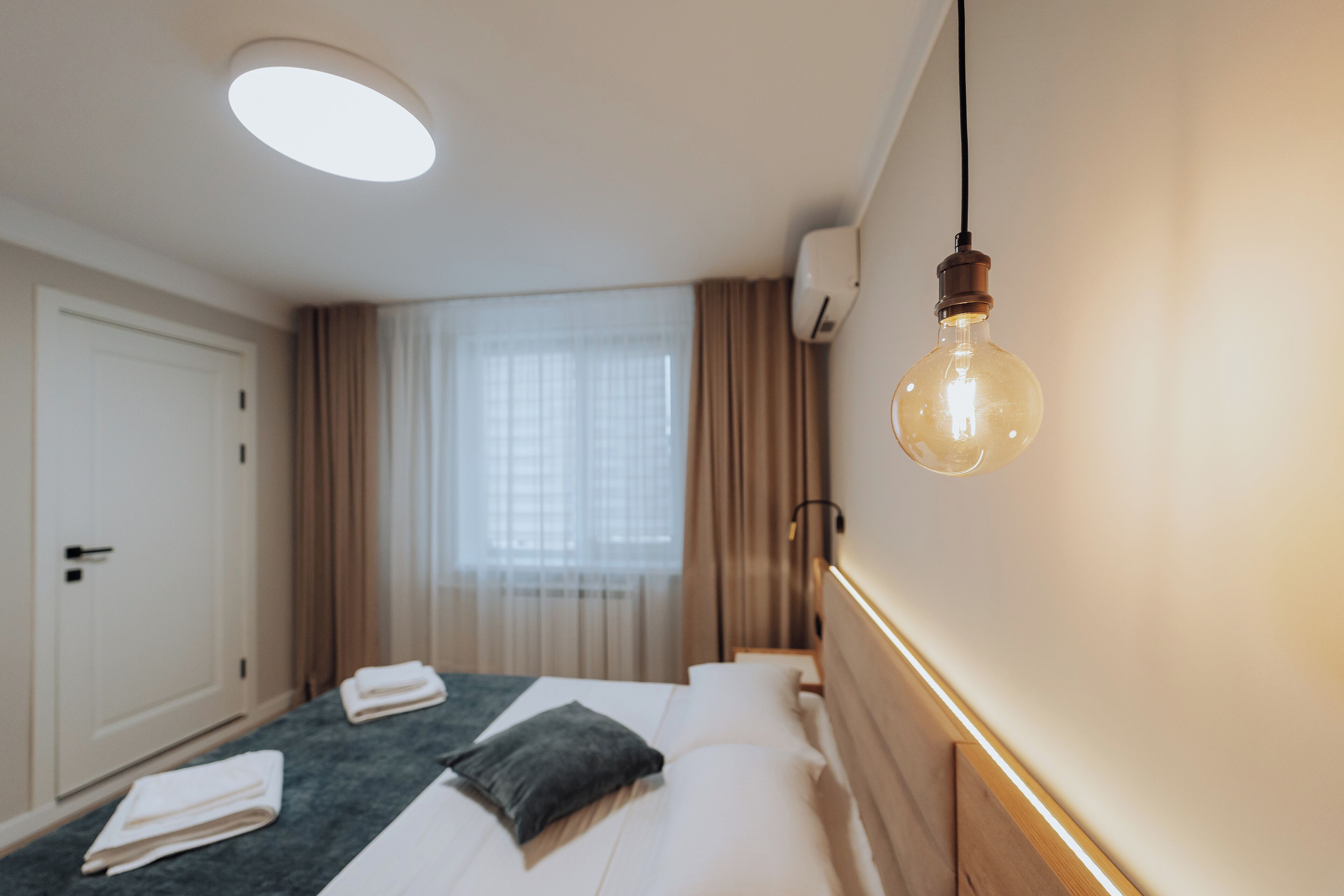 A hotel room with a bed, a bedside table, a hanging light bulb, and a window with curtains.