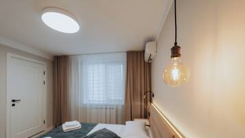 A hotel room with a bed, a bedside table, a hanging light bulb, and a window with curtains.