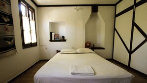 A neatly made bed with white linens in a room with wooden flooring, a window with curtains, and a wall-mounted shelf with books. - Secluded Cottage Guesthouse in the Mountains (Volos)