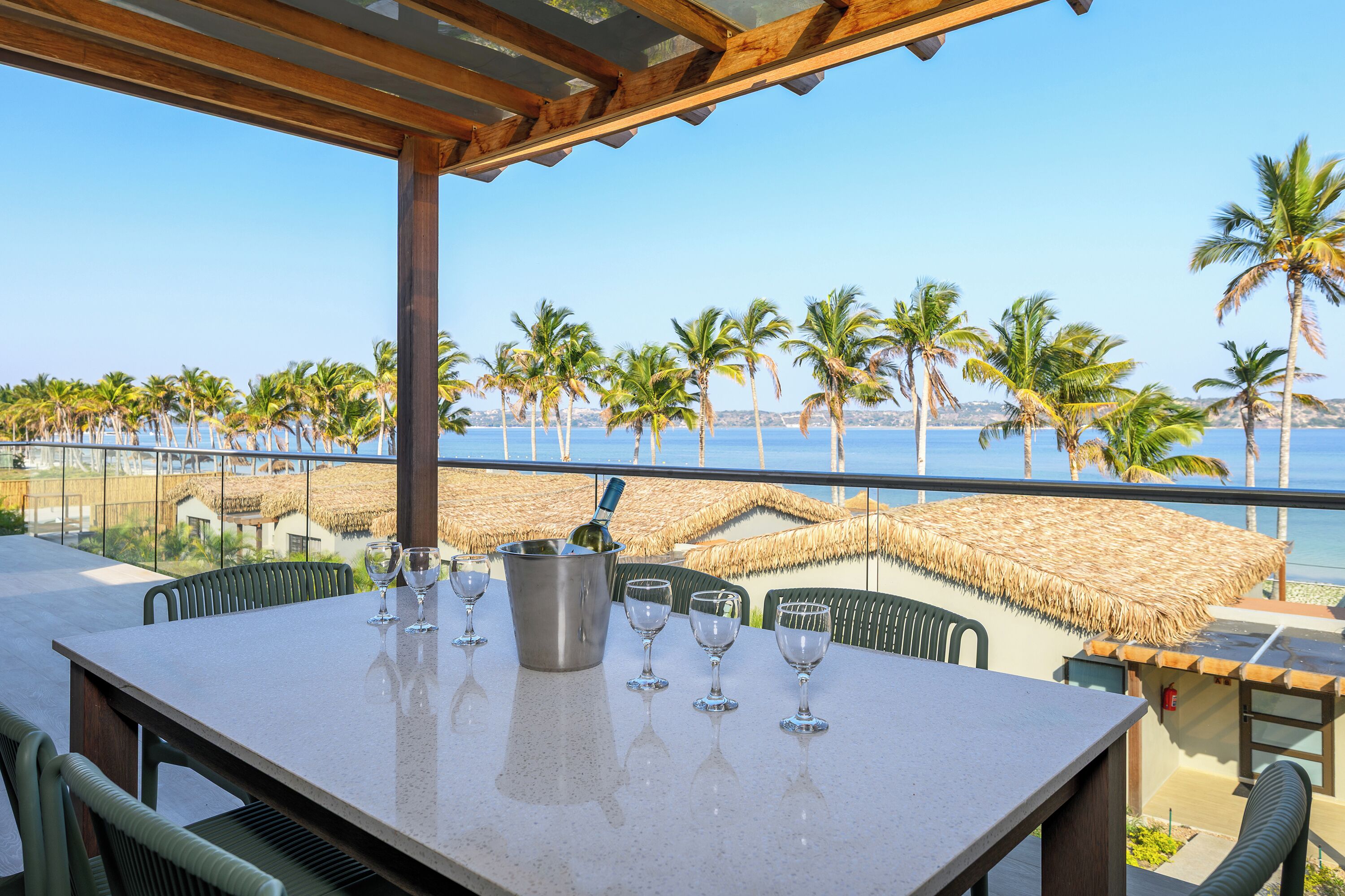 A terrace with a table set for dining, overlooking a beach with palm trees and the sea.