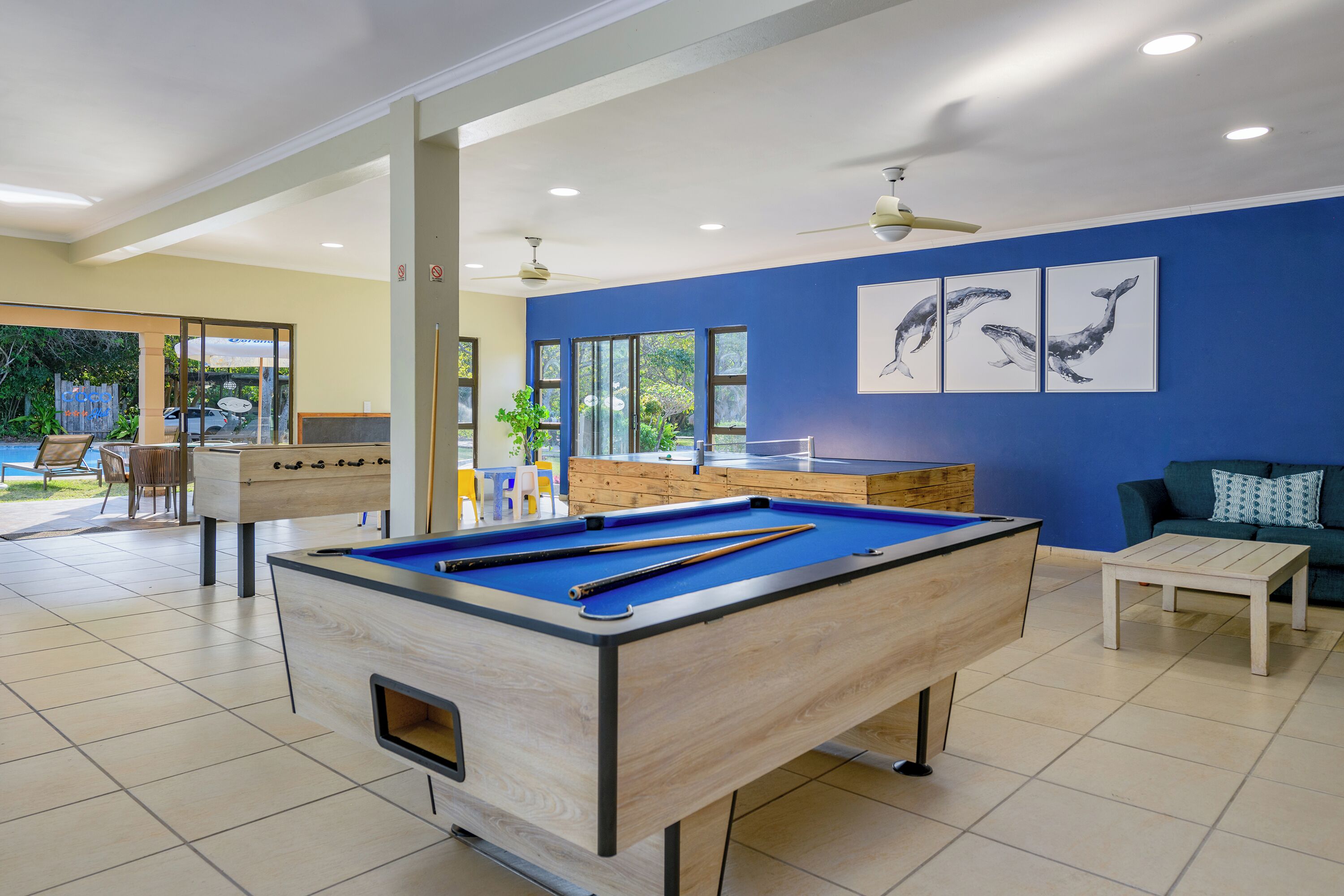 A pool table in a room with a blue wall, a ceiling fan, and a view of the outdoors.