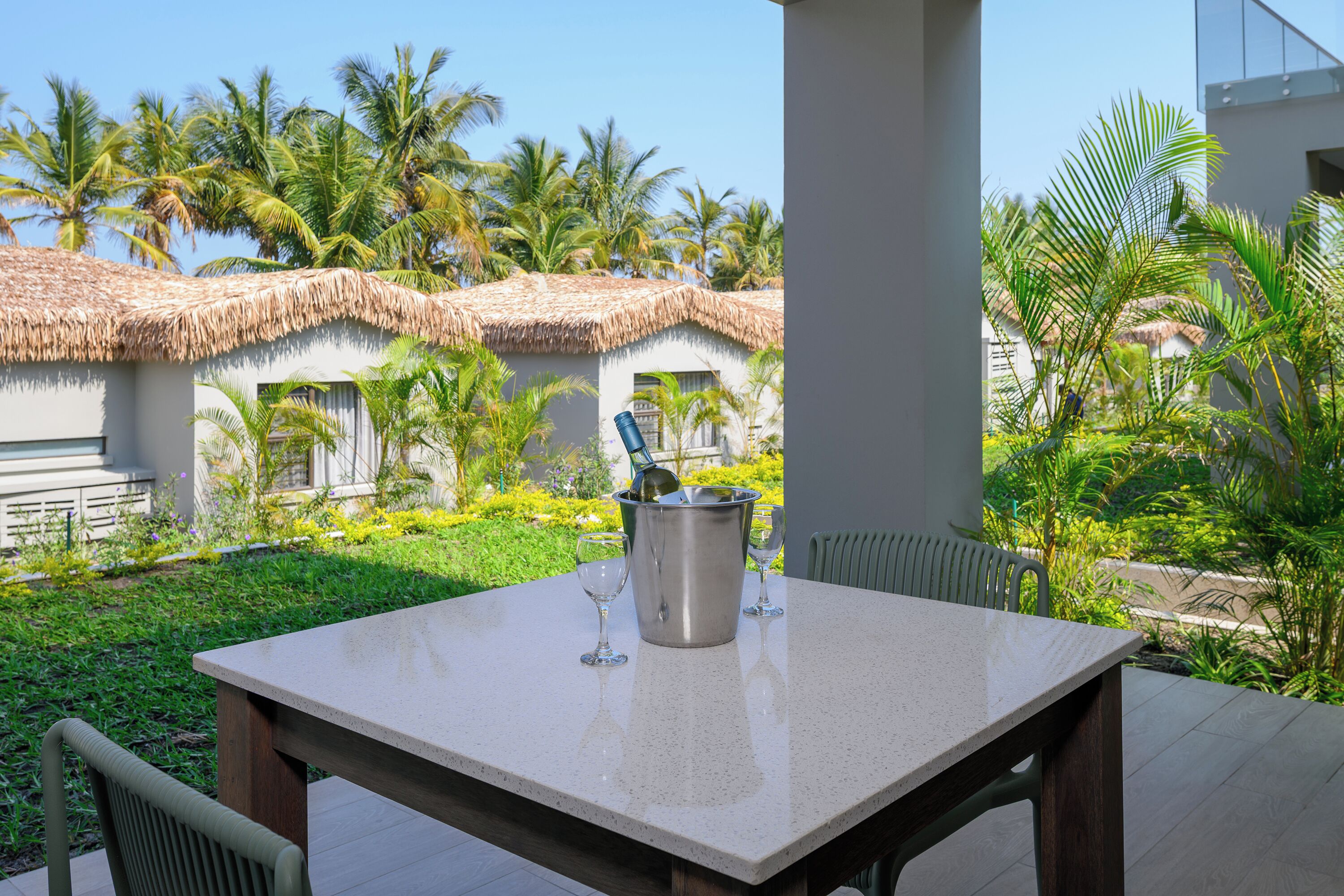 Un patio con una mesa, una copa de vino y un balde con una botella, rodeado de palmeras y un edificio con techo de paja.