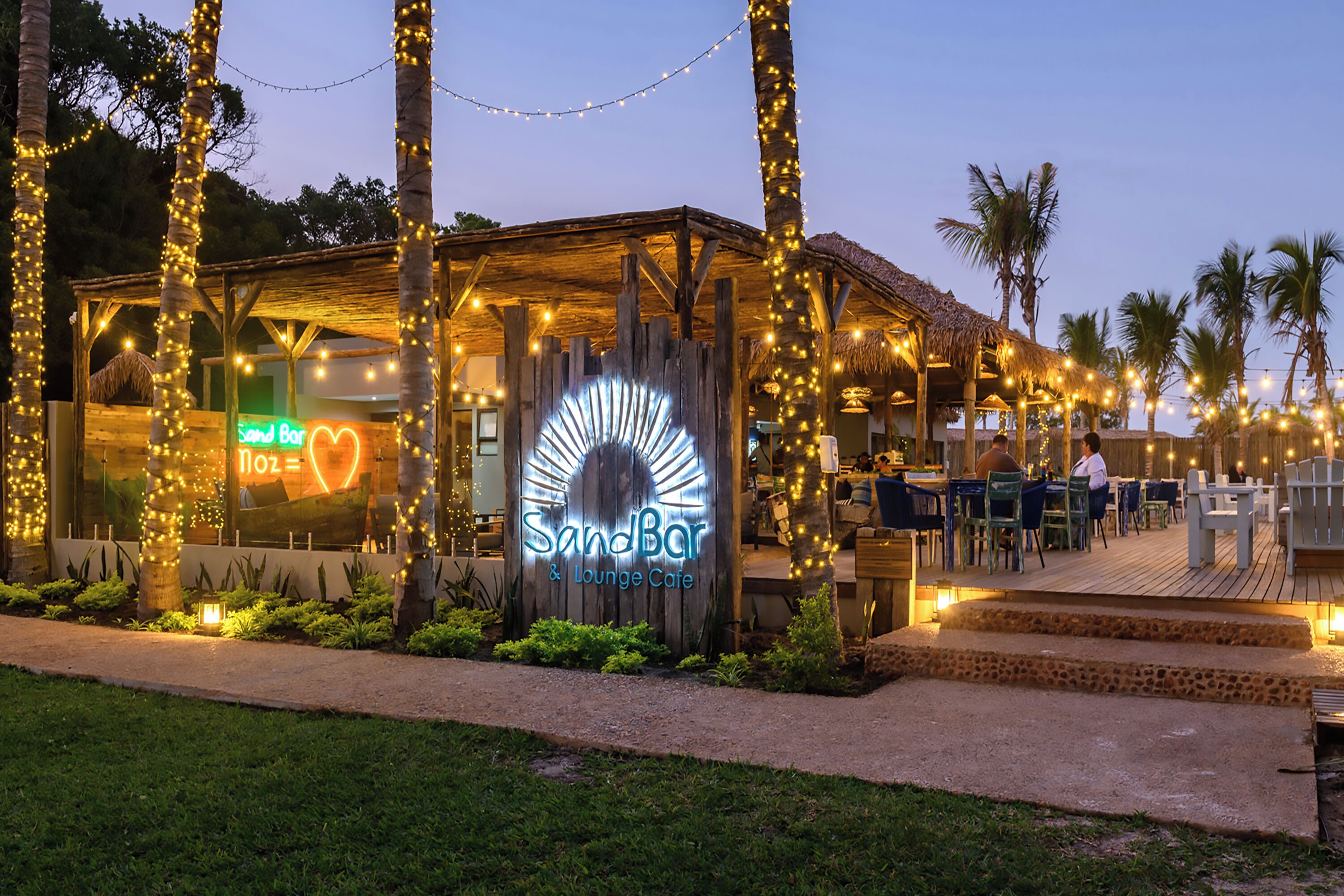 A beachside lounge with a wooden structure, string lights, and a neon sign.