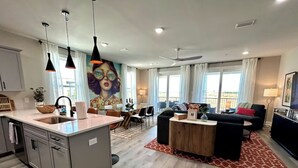 A modern kitchen with a central island, dining area with a table and chairs, a living room with a sofa and a TV, and large windows with curtains.
