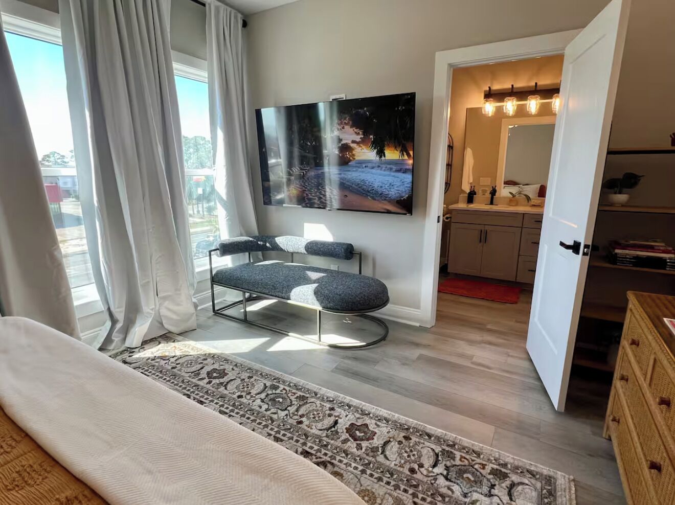 A bedroom with a bed, a bench, a large window with curtains, a TV, a mirror, and a door leading to a bathroom.