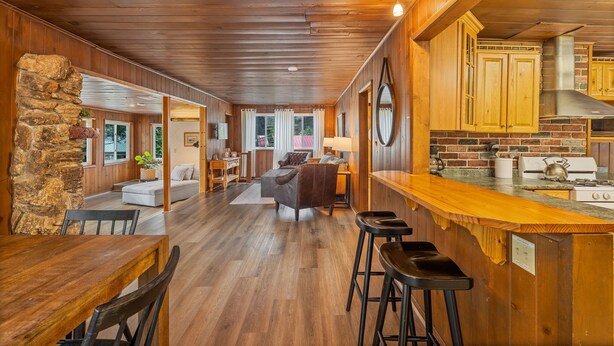 Une cuisine spacieuse avec des meubles en bois et un mur de crédence en briques, un coin repas avec une table et des chaises en bois, et un salon confortable doté d’une cheminée.