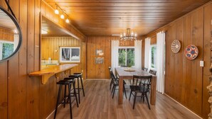 Un intérieur en bois comprenant une table à manger et des chaises, un coin bar avec des tabourets et une grande fenêtre avec des rideaux.