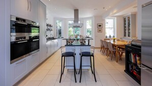 A modern kitchen with built-in appliances, a central island with bar stools, and a dining area with a wooden table and chairs. - The Kensington Townhouse, by Veeve (London)