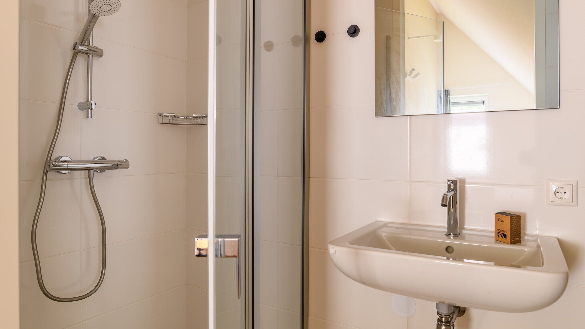 A bathroom with a glass shower enclosure, a sink with a mirror above it, and a small box labeled "DORMIO" on the wall.