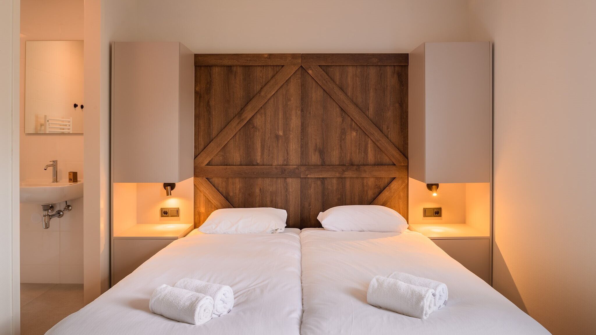 Una habitación de hotel con cabecera de madera, dos camas, lámparas de noche, un lavamanos y toallas.