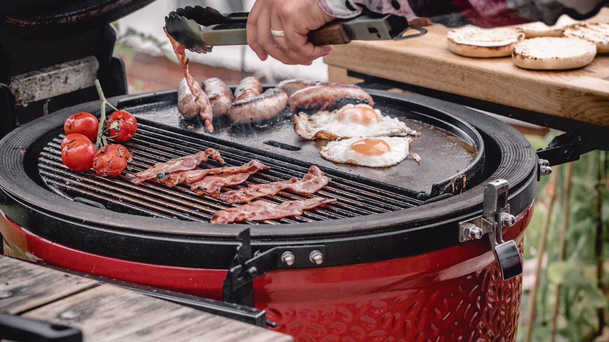 Een rode barbecue waarop spek, eieren en tomaten worden gebakken.