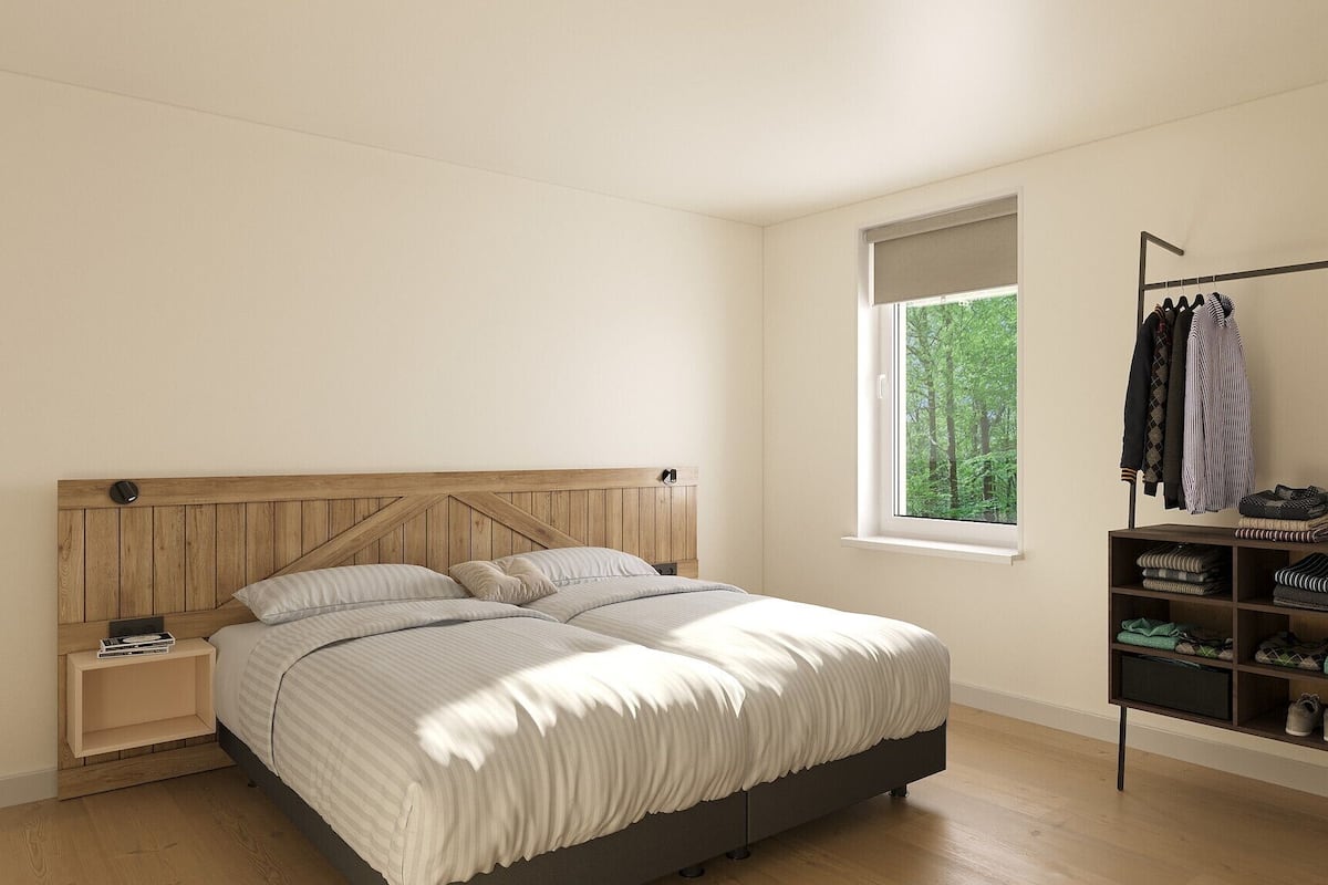 A bedroom with a wooden headboard, a bed with striped bedding, a nightstand, a window with a view of greenery, and a clothing rack with folded clothes and shoes.