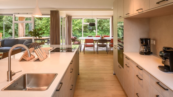 A modern kitchen with a central island, stainless steel sink, and wooden cabinets. There is a dining area with a table and chairs, and large windows offering a view of greenery. - Dormio Resort De Hondsrug (Gasselte)