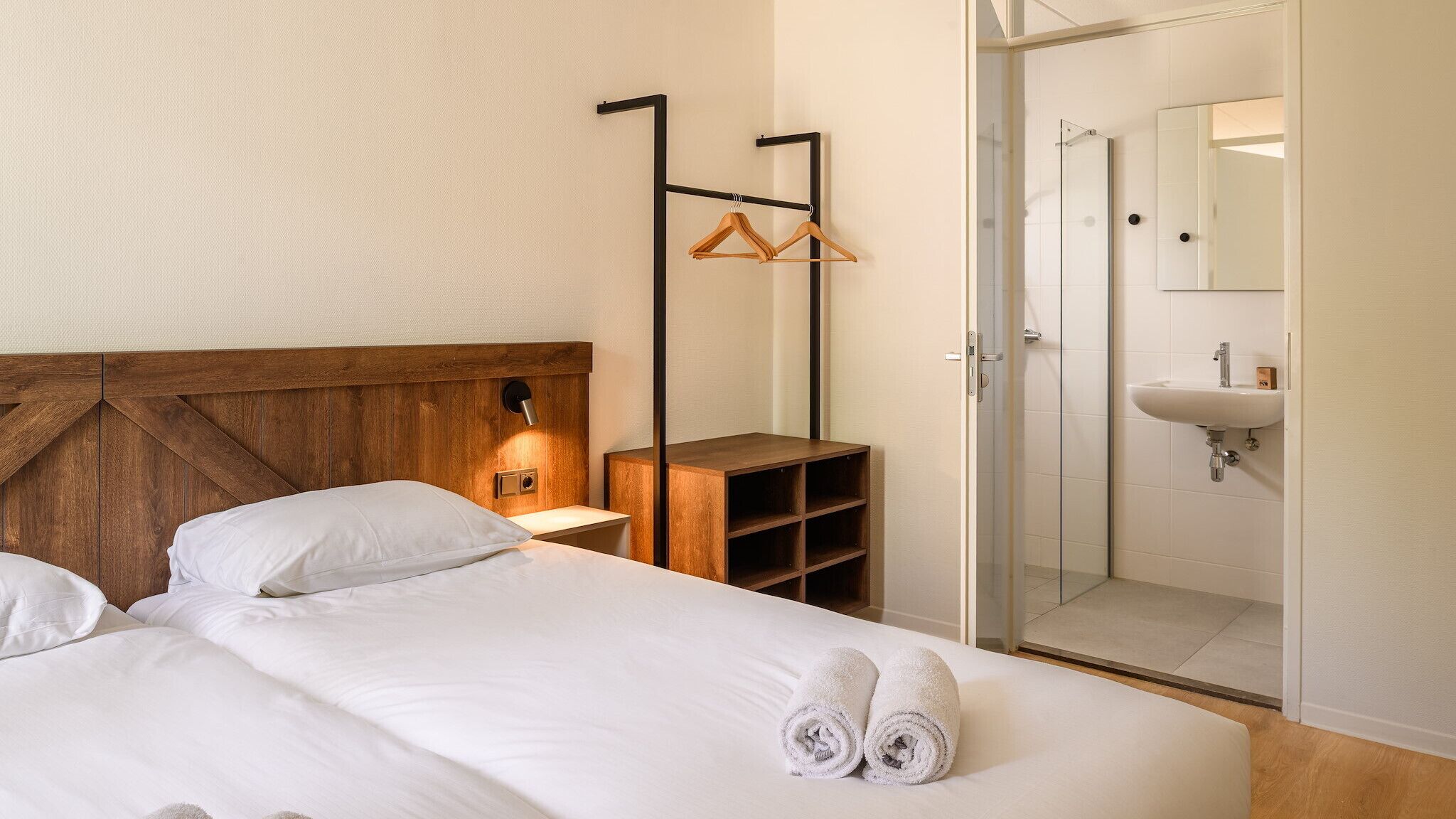 Ein Hotelzimmer mit einem hölzernen Kopfteil, einem Nachttisch mit Lampe, einem Kleiderschrank und einem Badezimmer mit Waschbecken und Spiegel.