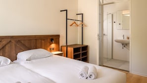 Habitación de hotel con cabecera de madera, mesita de noche con lámpara, armario y baño con lavabo y espejo.