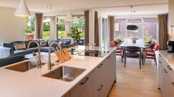 Una cocina moderna con una isla central, fregaderos de acero inoxidable y un comedor con una mesa larga y sillas.