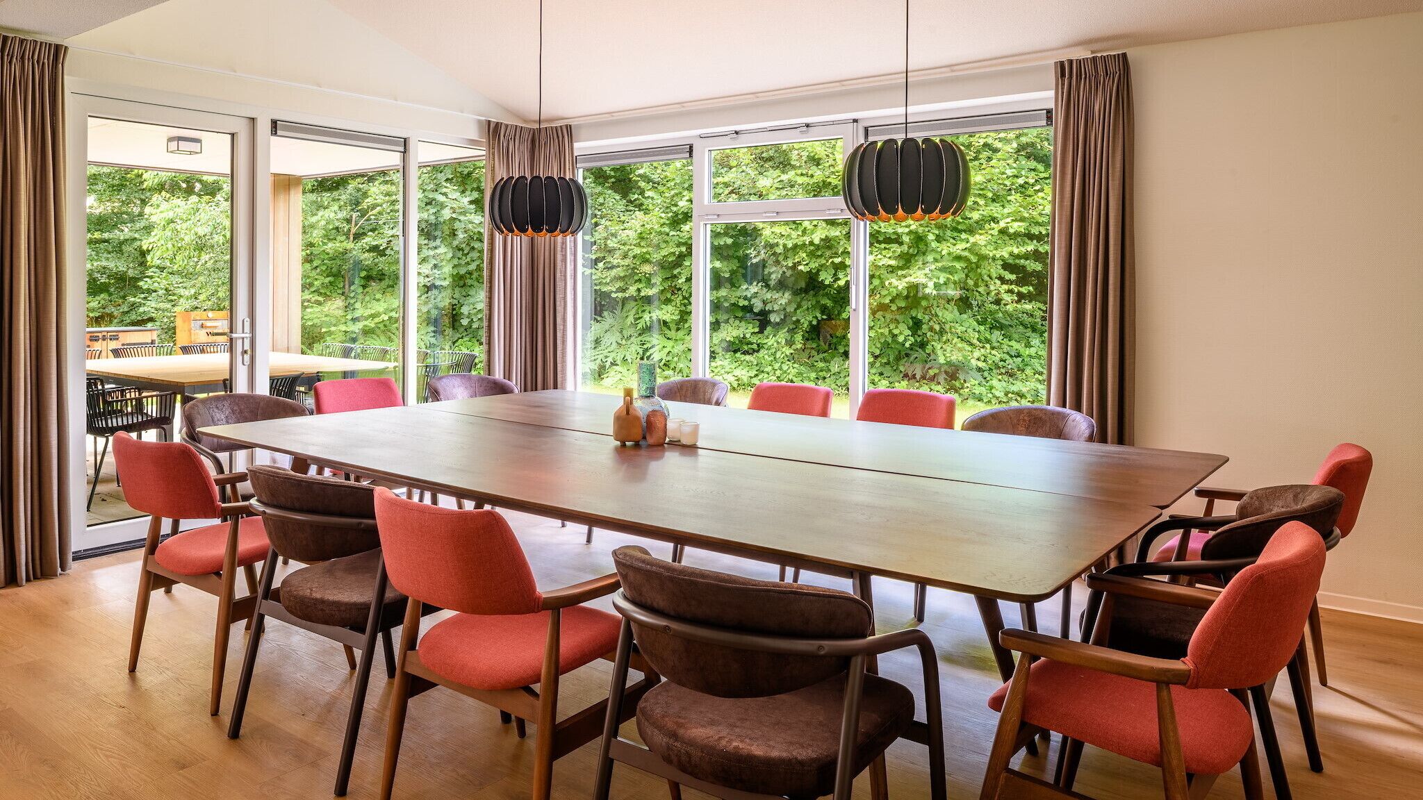 A conference room with a long table and multiple chairs, large windows with curtains, and hanging pendant lights.