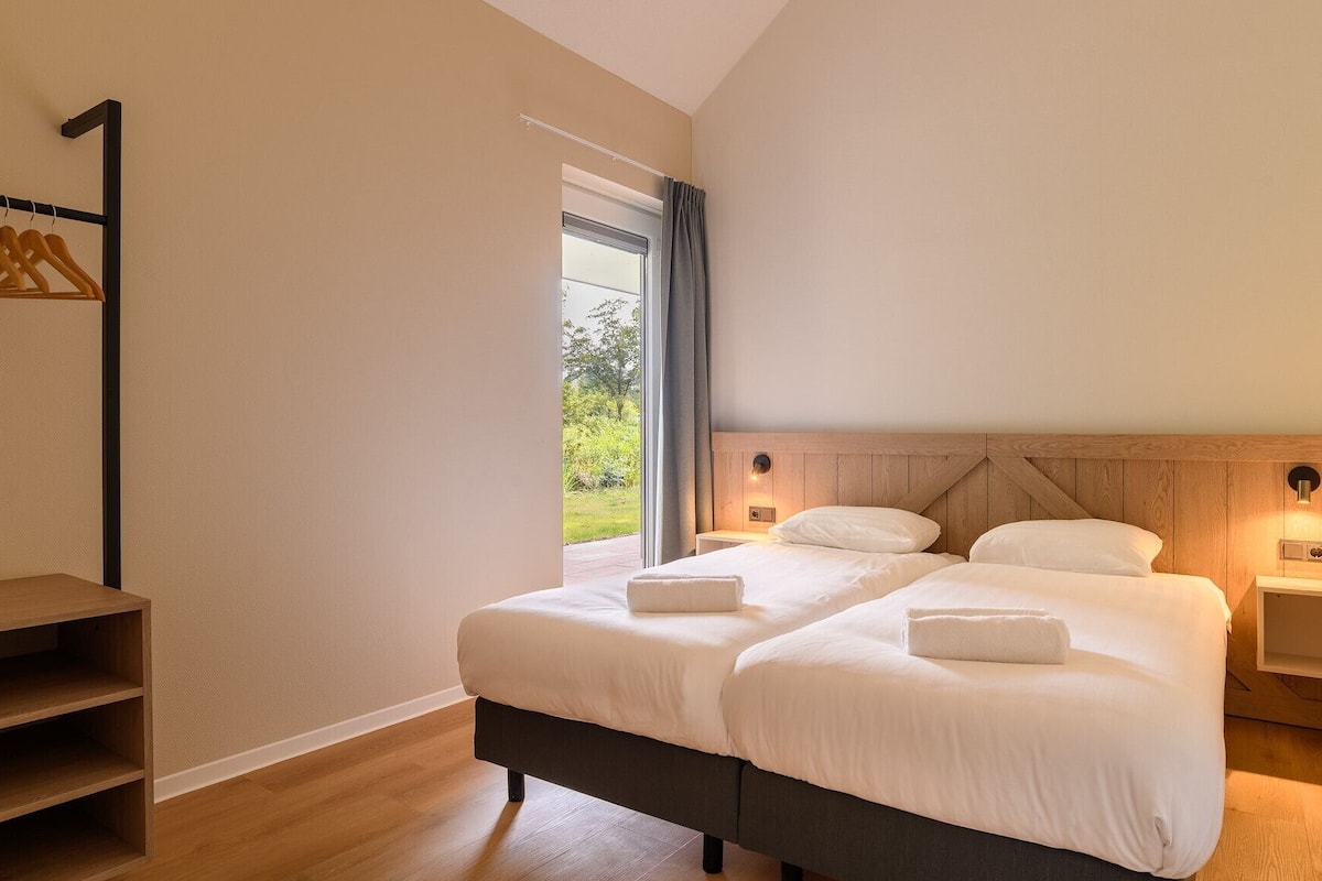 A bedroom with two beds, a wooden headboard, bedside tables, a wooden shelf, and a window with a view of greenery.