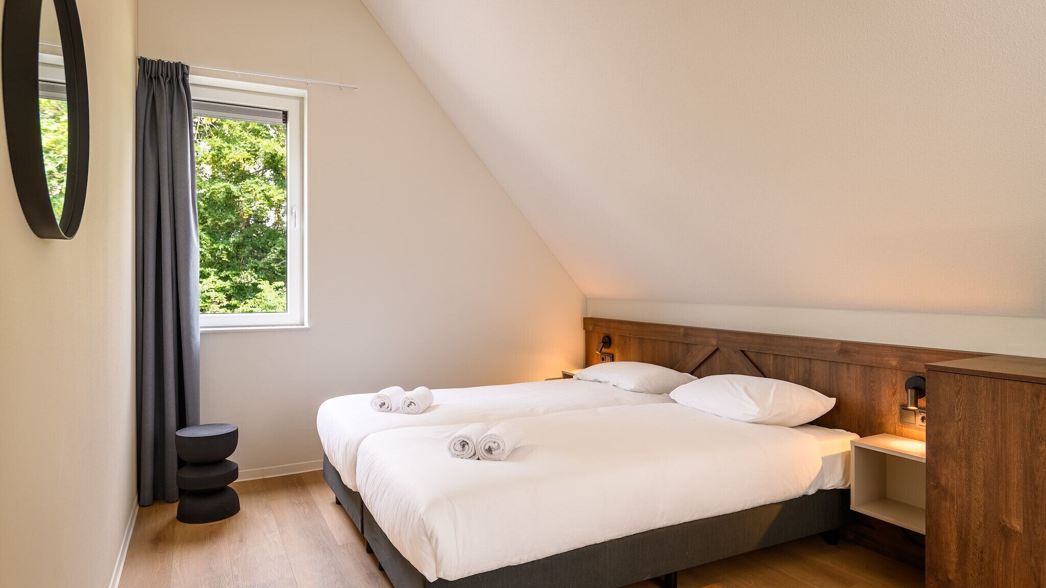 Une chambre avec deux lits, une fenêtre avec des rideaux, une table de chevet avec une lampe et un petit tabouret noir.