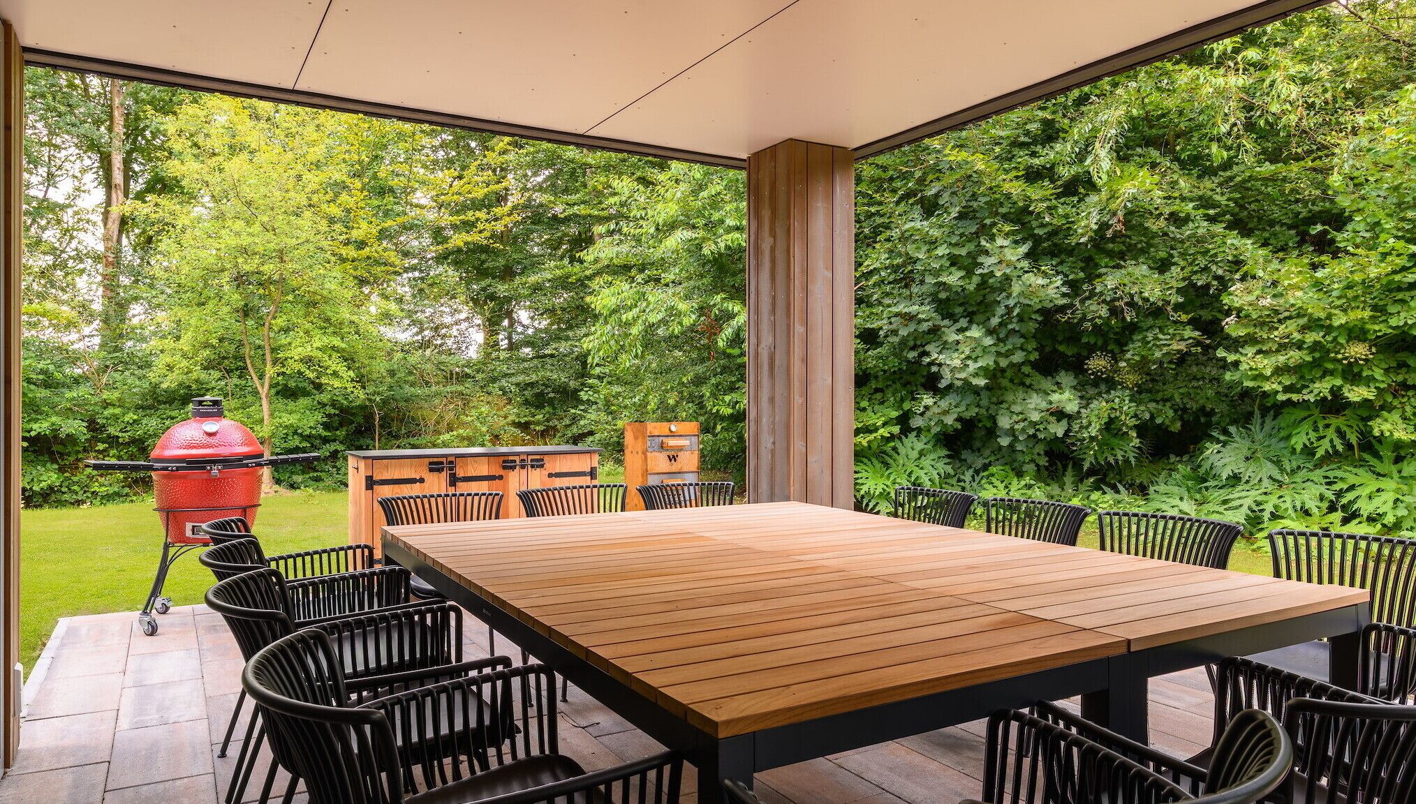 Een ruim terras met een houten tafel en stoelen, een rode barbecue en een houten kast.