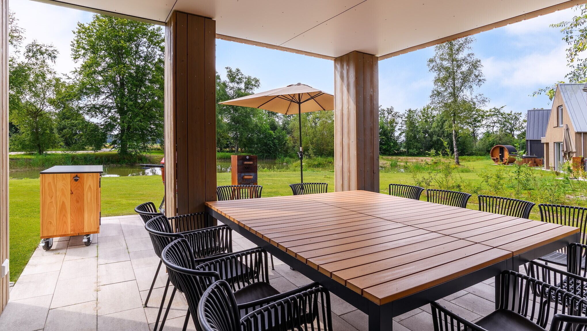 Eine überdachte Terrasse mit Holztisch und -stühlen, einem Holzschrank und einem Sonnenschirm.