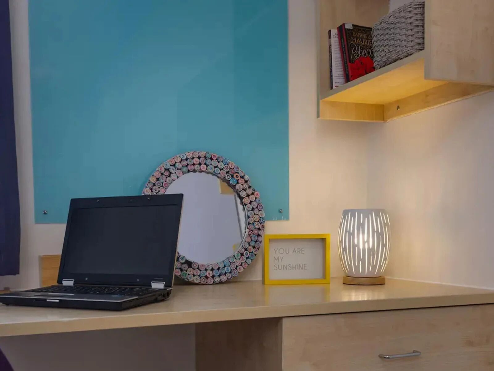 A desk with a laptop, a framed quote, a decorative mirror, and a lamp.