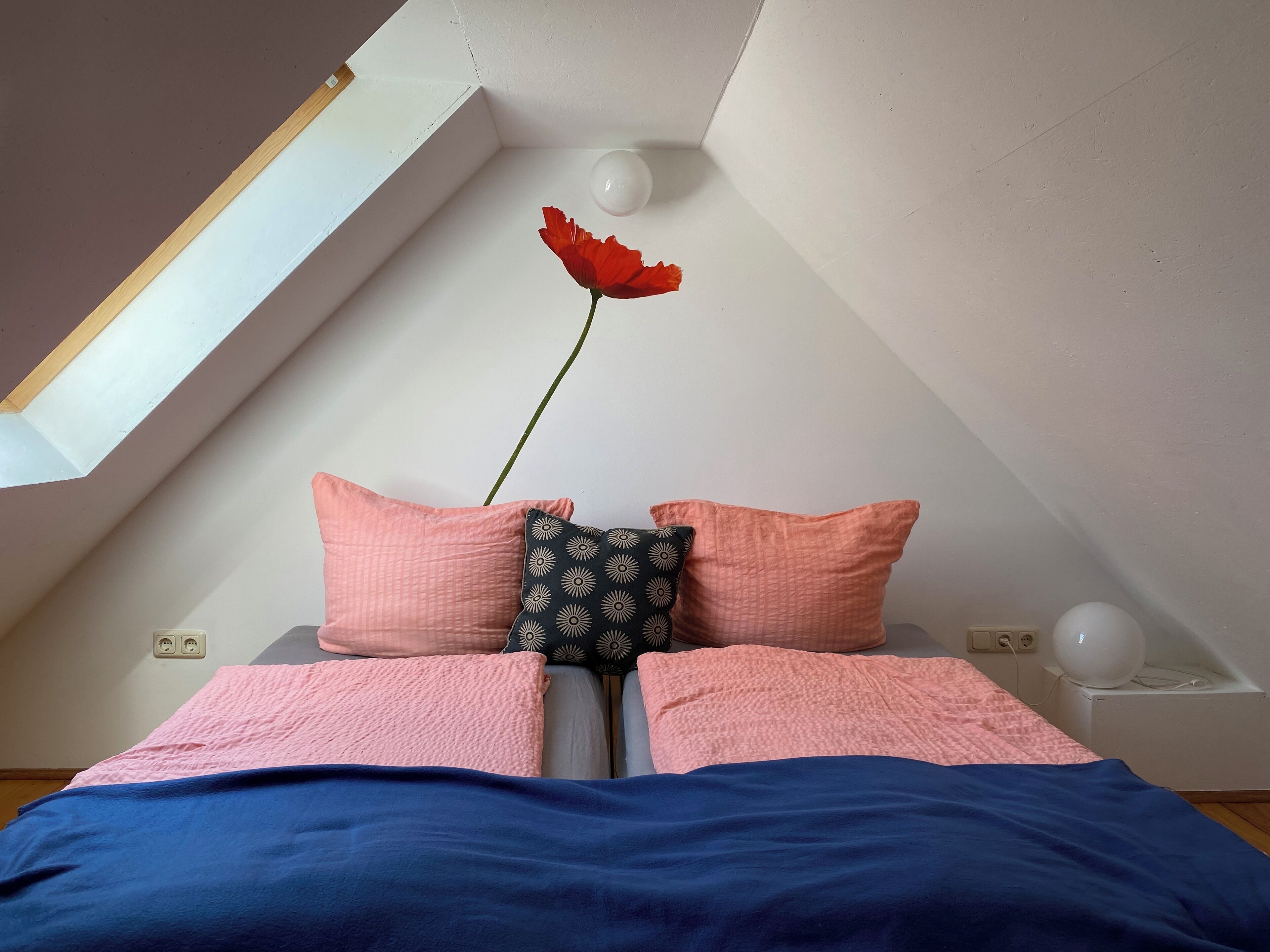 Een slaapkamer met een schuine plafond, een bed met roze en blauwe beddengoed, twee kussens, en een rode bloem aan de muur.