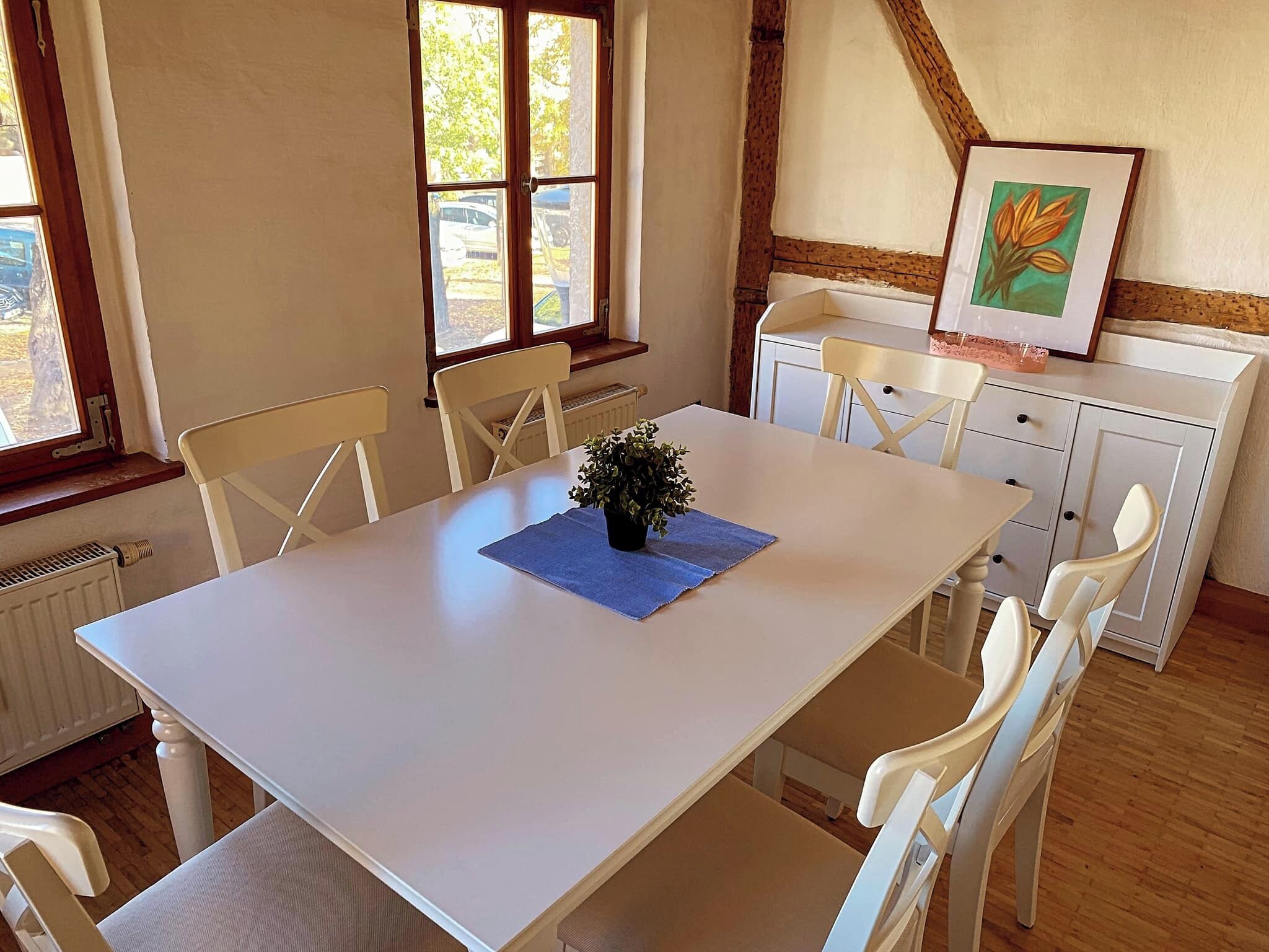 A dining area with a white table, four white chairs, a potted plant, and a framed picture on a cabinet.