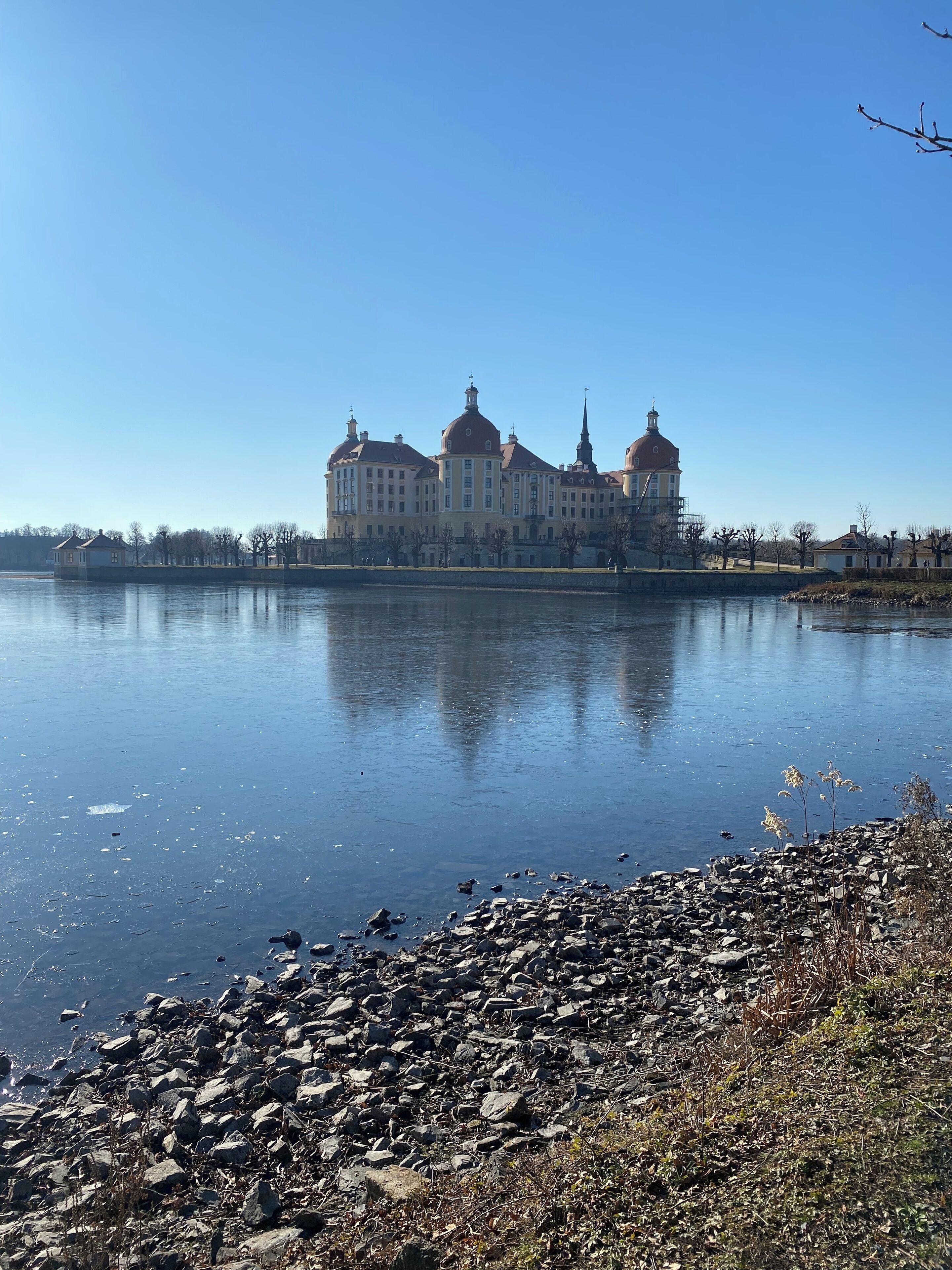 A large, historic building with multiple domes and a spire, situated by a calm body of water.