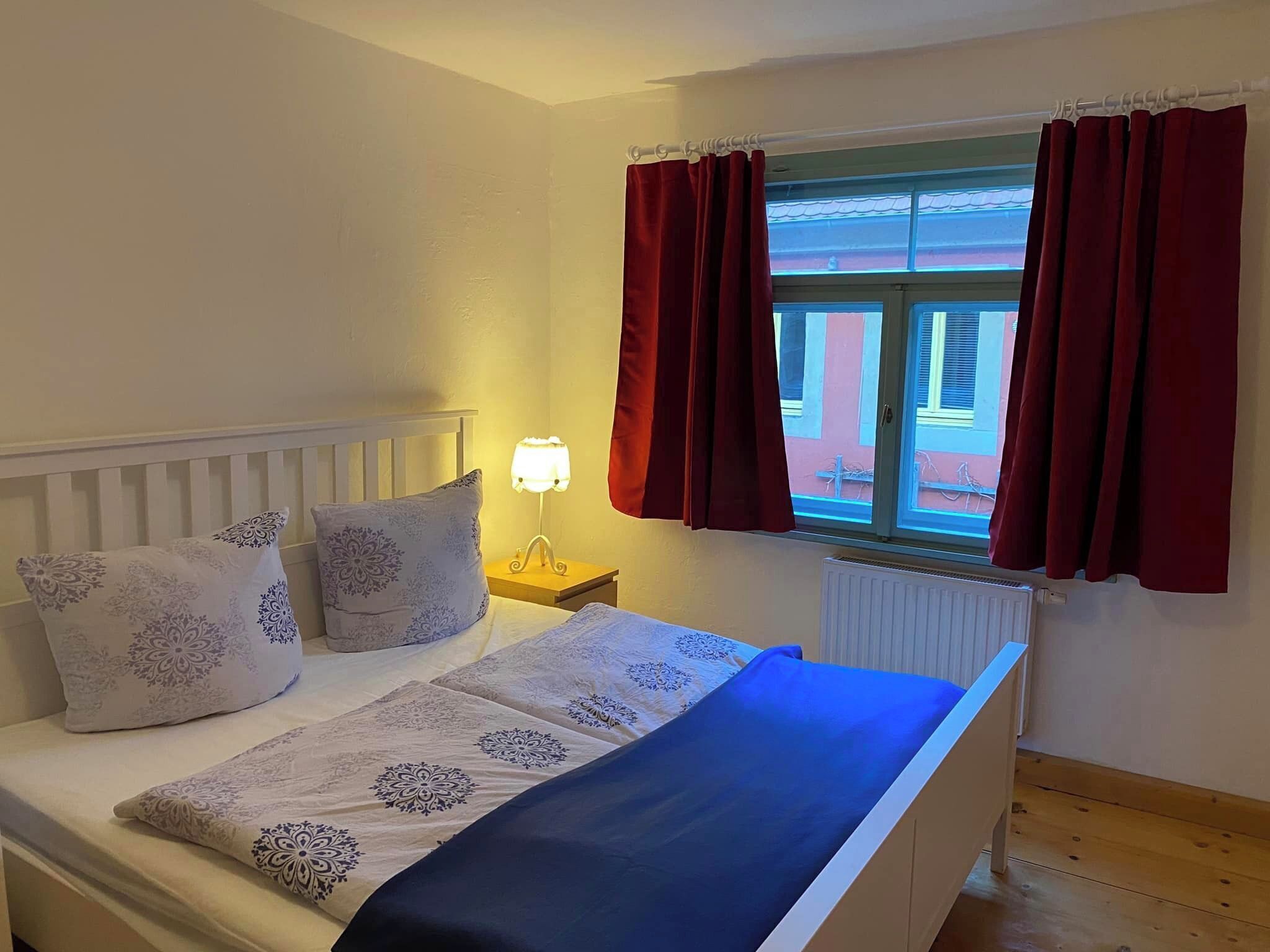 Ein Schlafzimmer mit Bett, Nachttisch, Lampe und einem Fenster mit Vorhängen.
