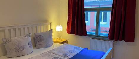 Ein Schlafzimmer mit Bett, Nachttisch, Lampe und einem Fenster mit Vorhängen.