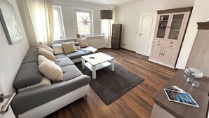 A modern living room with a grey sectional sofa, a white coffee table, and a wooden floor. There is a door with glass panels and a small table with magazines.