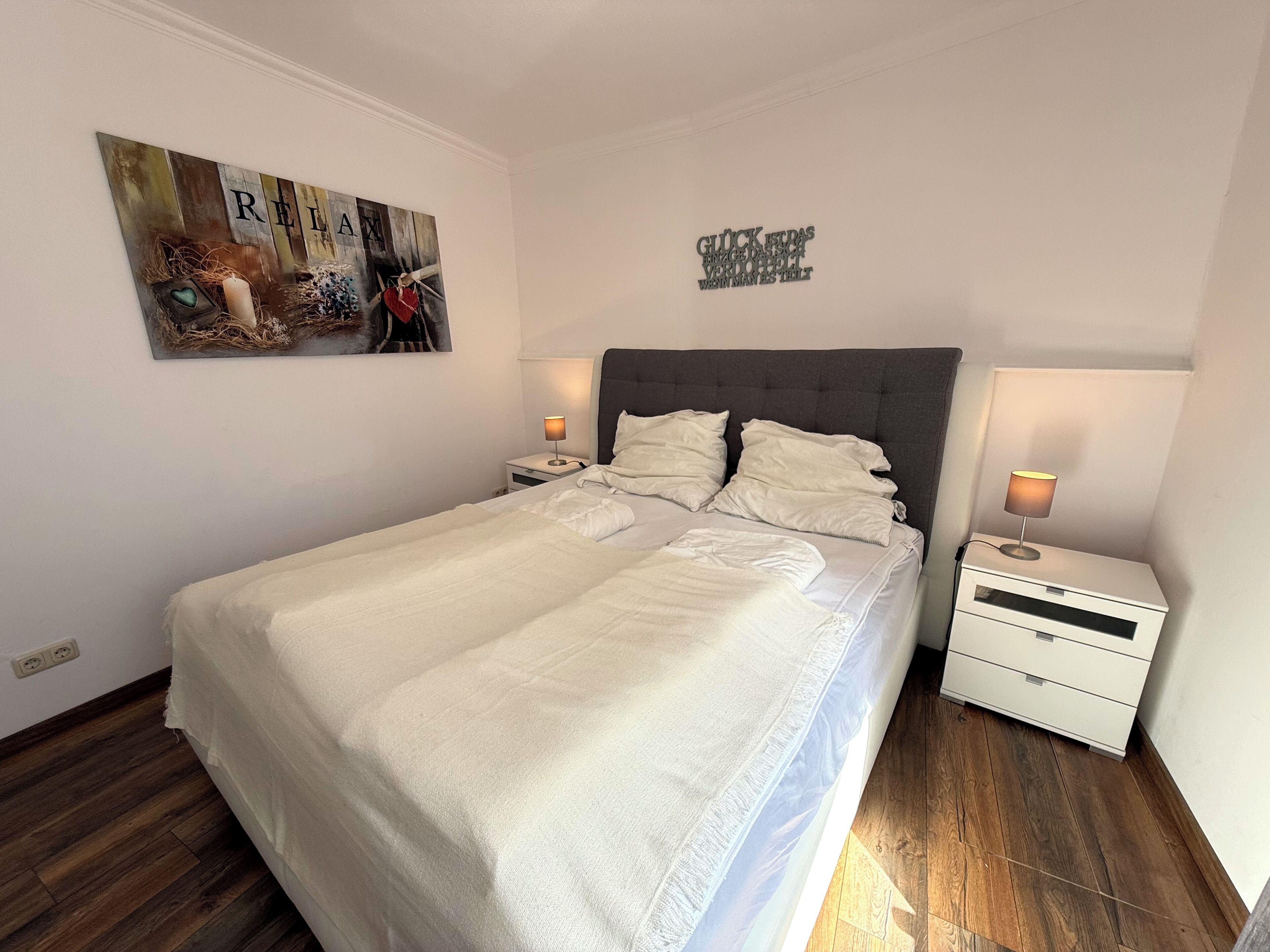 Un lit bien fait, avec du linge de lit blanc, une tête de lit rembourrée grise et deux tables de chevet avec des lampes. Un tableau sur lequel est écrit le mot « RELIANCE » est accroché au mur.