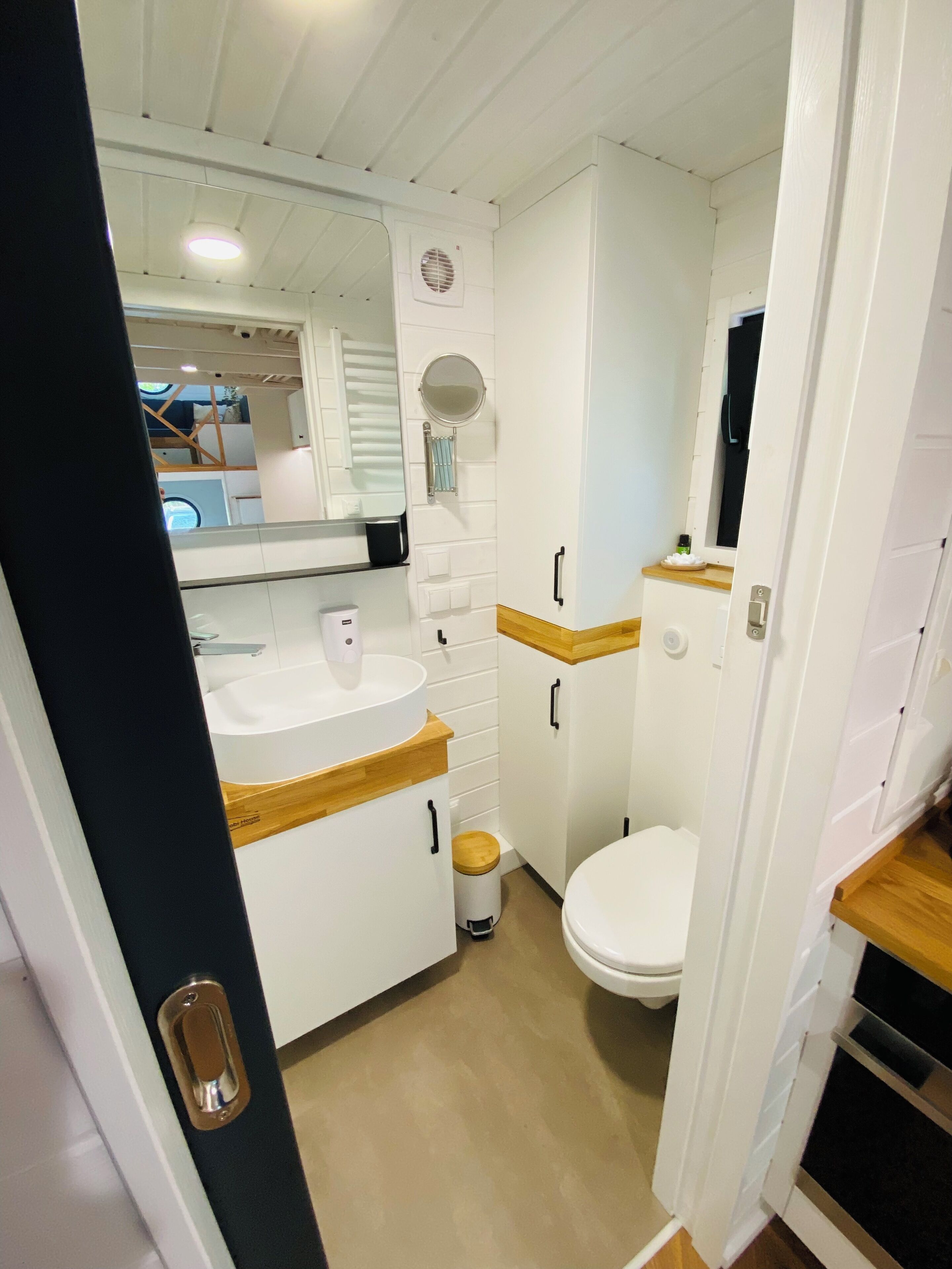 A compact, white bathroom with a sink, mirror, and toilet. It features wooden accents on the countertops and cabinets.