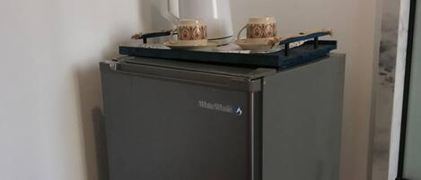 A compact, dark-colored refrigerator with a brand label, placed in a room with wooden flooring and a red bed.