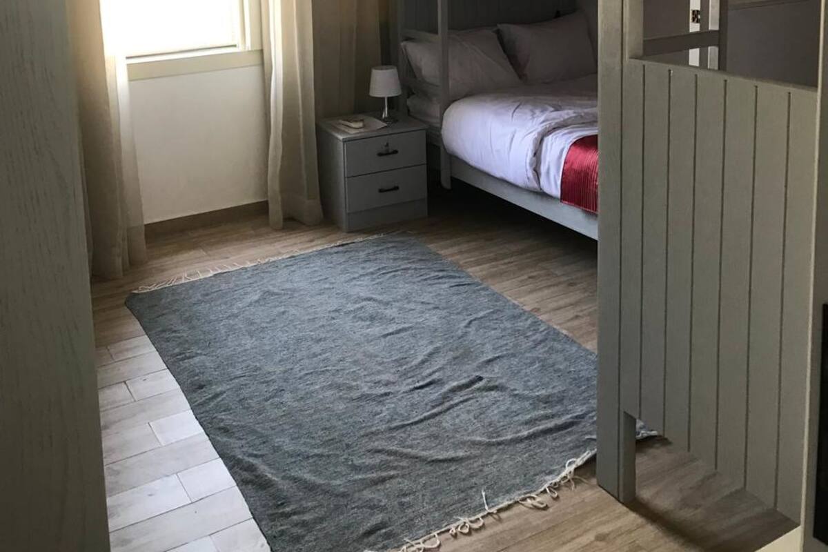 Un dormitorio con una cama litera, una mesita de noche con lámpara, una alfombra gris y un armario.