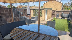 A covered outdoor patio with a round table and chairs, a wooden deck, and a brick wall in the background. - Modern 3BR Family Home w/ Garden | Luton (Luton)