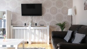 A living room with a flat-screen TV mounted on the wall, a white radiator, a dark brown sofa, and patterned wallpaper. - Modern 3BR Family Home w/ Garden | Luton (Luton)