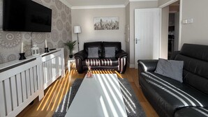 A modern living room with a black leather sofa, a white coffee table, and a TV mounted on the wall. - Modern 3BR Family Home w/ Garden | Luton (Luton)