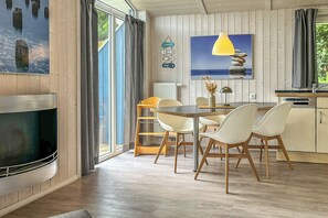 A modern kitchen with wooden cabinets, a dining area with white chairs, a TV, and a large window with curtains.