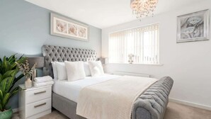 A bedroom with a tufted headboard, white bedding, a bedside plant, and artwork on the walls.