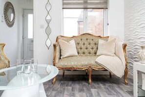 Une pièce avec un canapé capitonné, une table en verre avec des verres, un miroir et un mur orné d’un motif.