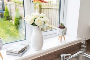 Un vase blanc orné de fleurs blanches, posé sur un rebord de fenêtre, une petite plante en pot et une pile de livres.