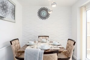 Comedor con una mesa redonda dispuesta para cuatro personas, un reloj de pared decorativo e iluminación colgante.