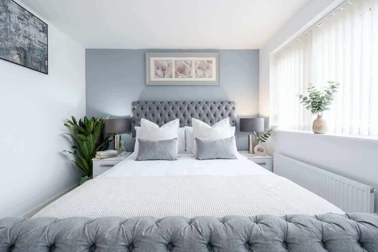 Une chambre à coucher avec un grand lit, une tête de lit capitonnée, des tables de chevet avec des lampes et une fenêtre avec des rideaux.