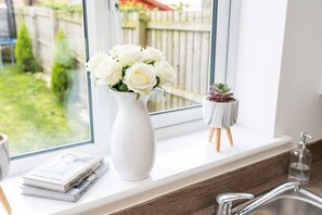 Un jarrón blanco con flores blancas en un alféizar, una planta pequeña en maceta y una pila de libros.