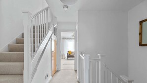 A hallway with a carpeted staircase, white walls, and a door leading to a room with a bed and a window. - Central Exeter 3BR | Walk Everywhere (Exeter)