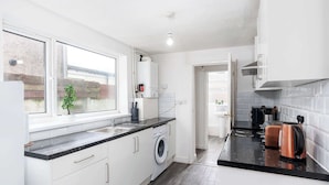 A modern kitchen with white cabinets, a built-in washing machine, a black countertop, and a view of the outside through a window. - Stylish 4BR Family Home | Heart of Grimsby (Grimsby)