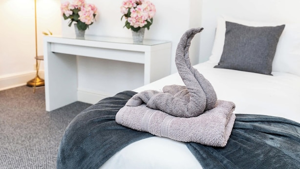A neatly made bed with a folded towel in the shape of a swan, a white bedside table with two vases of flowers, and a grey pillow on the bed. - Stylish 4BR Family Home | Heart of Grimsby (Grimsby)