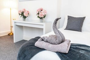 Un lit bien fait, avec une serviette pliée en forme de cygne, une table de chevet blanche ornée de deux vases de fleurs et un oreiller gris sur le lit.